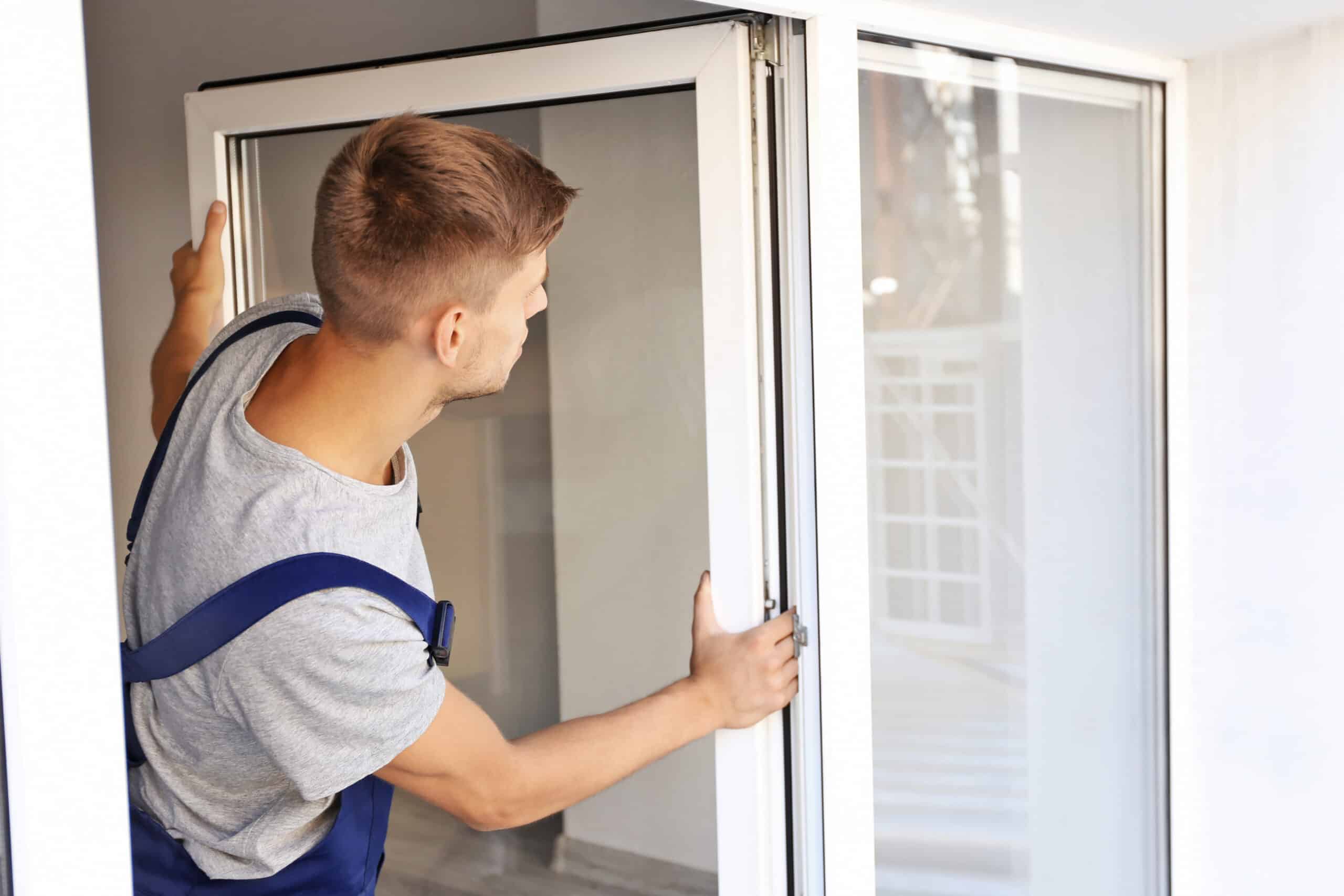 A worker in a gray shirt and overalls installs a sliding glass door in a bright, contemporary room.