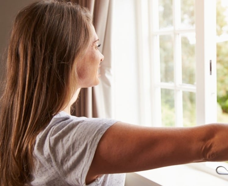 Woman in a light t-shirt looking out a window with her left arm extended, in a brightly lit room, capturing the essence of how to brighten your home for spring.