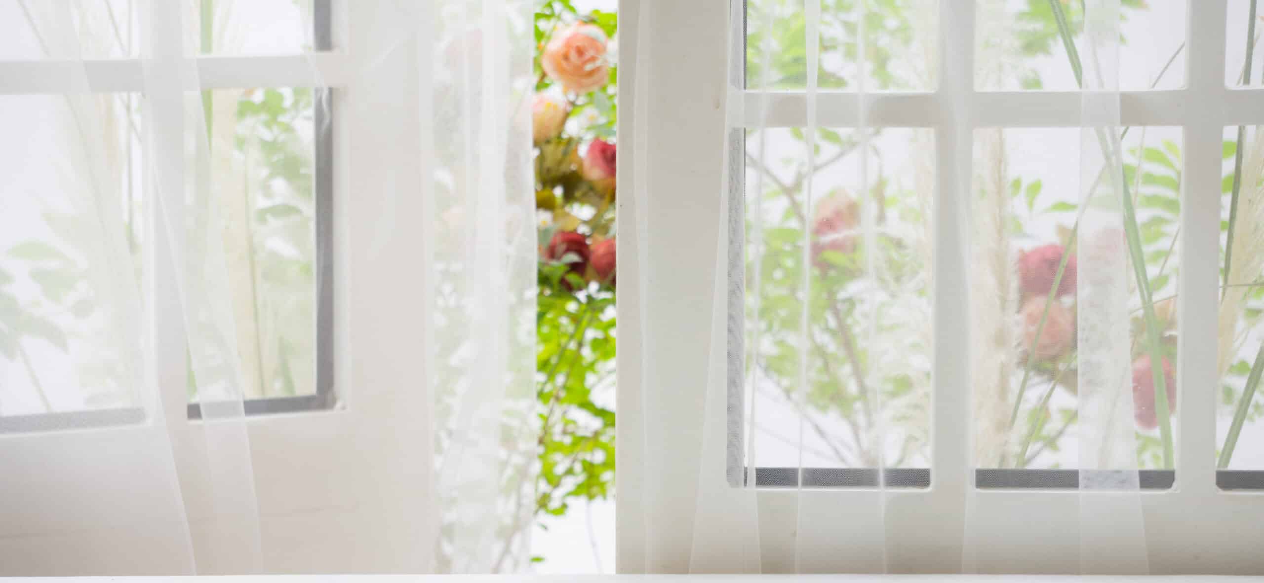 Sheer white curtains billow gently in a breezy room with a view of a garden through an energy-efficient windowpane, showing glimpses of green leaves and pink flowers.