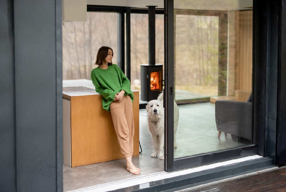 A woman in a green sweater and brown pants leans on a wooden counter by an open door, looking outside through a new window chosen from a top-rated window company, where a white dog stands on the