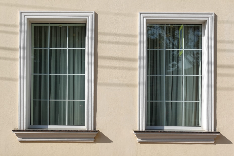 Two framed windows with closed vertical blinds on a beige wall, one window reflecting sunlight, installed by a reputed window company.