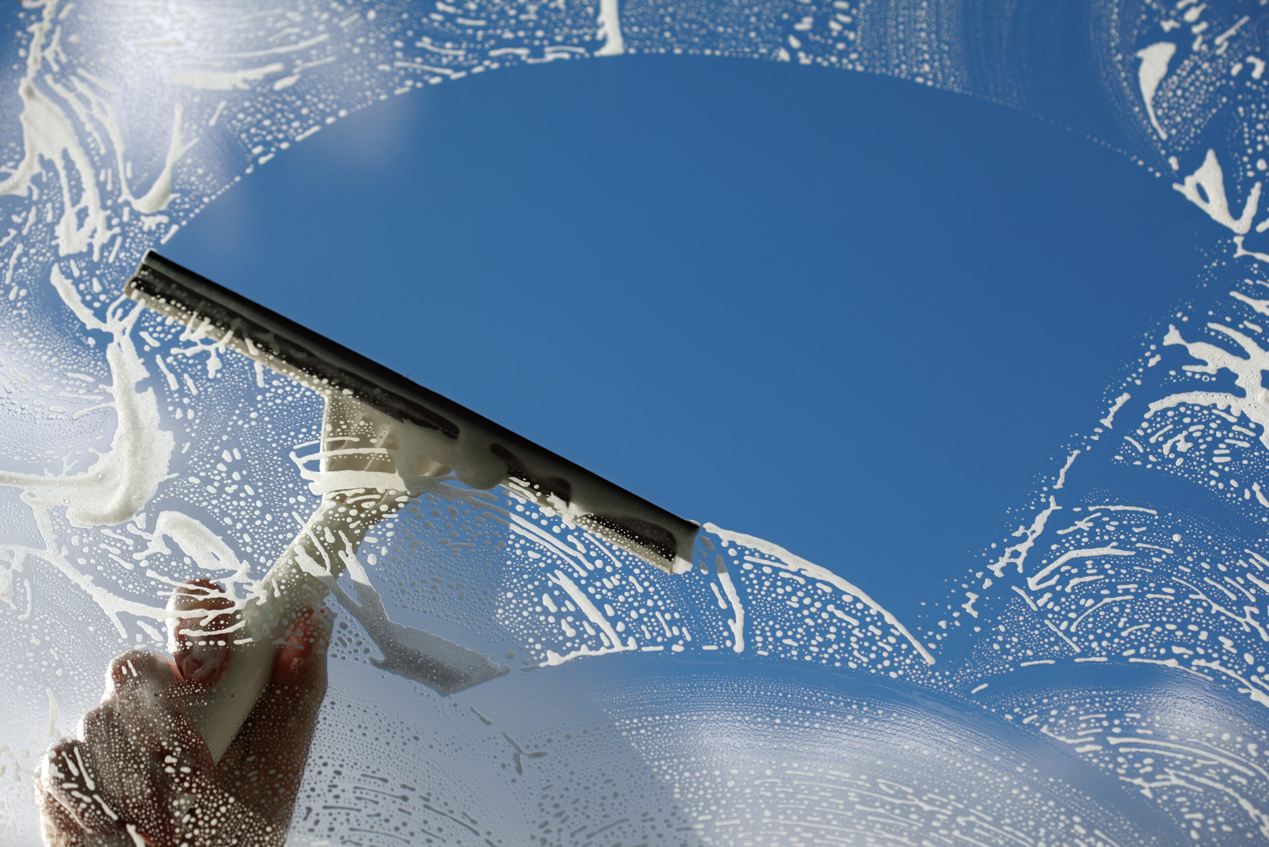 Hand using a squeegee for spring cleaning, with soap suds visible across the window against a clear blue sky.