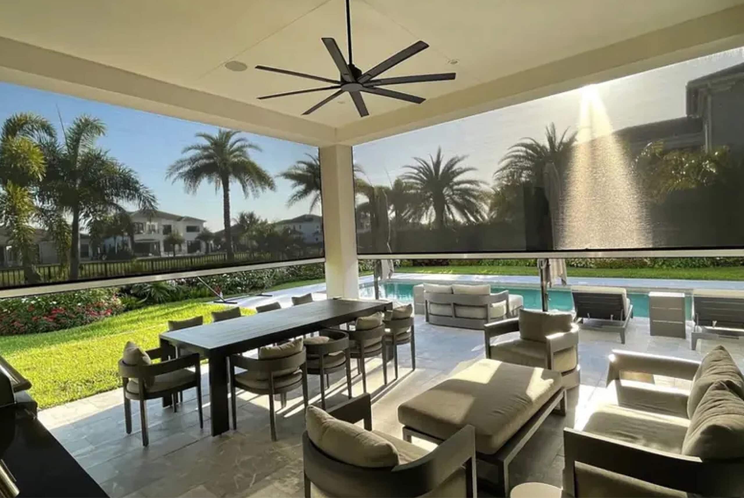 A covered patio with furniture, a ceiling fan, and retractable screens overlooks a swimming pool and landscaped yard in the background, all safeguarded by impact windows for added hurricane protection.